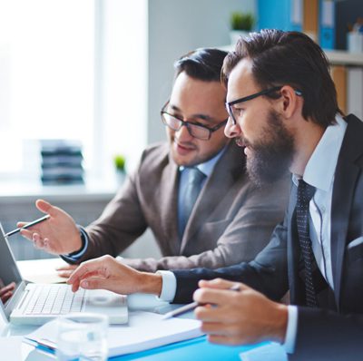 Two men in suits are sitting at a desk in an office setting. They are focused on a laptop, with one pointing at the screen and the other typing. Papers and a pen are on the table, and shelves with files are visible in the background.