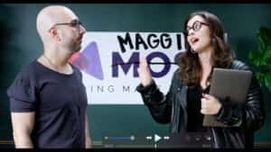 Progressive robot, two people talking in front of a “MAGGI MOS” sign, one holding a tablet and gesturing.