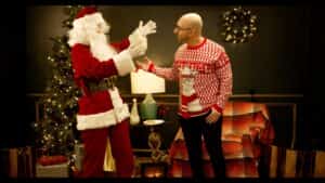 Progressive robot Santa Claus gestures while talking to a man in a red holiday sweater in a decorated Christmas room.