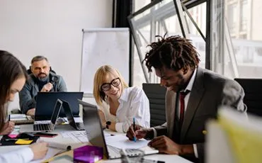Home - IT Consulting & Software Development 73 A group of people in a meeting room, sitting around a table with laptops and documents. A man in a suit writes on a paper, while a woman in glasses smiles at him. Others in the background are listening and working.