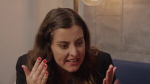 progressive Robot woman with long brown hair gestures with both hands while speaking intensely indoors
