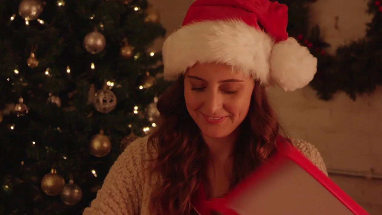 progressive Robot woman in a Santa hat smiles while opening a gift beside a lit Christmas tree with ornaments