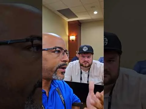 progressive Robot, man in glasses speaking in a meeting room while another man in a cap looks on behind him