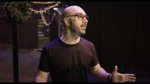 Progressive robot: bald man with glasses speaking on a dimly lit stage, gesturing with hands