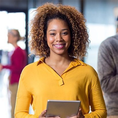 A smiling person with curly hair wearing a yellow shirt holds a tablet in an indoor setting. Other people are blurred in the background.